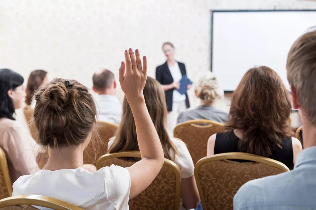 View of conference room meeting from behind woman with hair in but who is raising hand. 